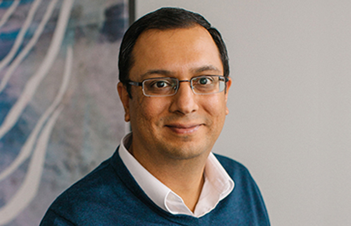 A professional individual stands confidently in an office environment, dressed in a smart blue sweater