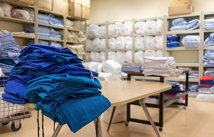 A neatly organized hospital linen room with stacks of folded blue scrubs on a table