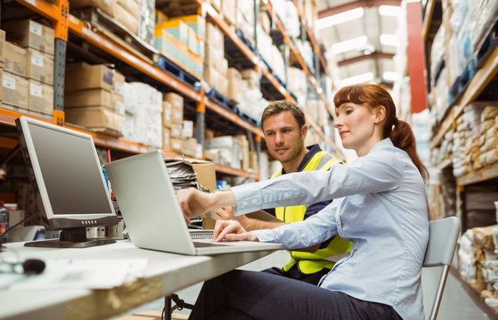 Two warehouse employees are engaged in a collaborative task at a workstation amidst the aisles