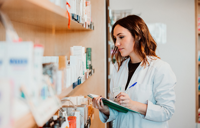 A female pharmacist in a white coat is attentively checking inventory and taking notes in