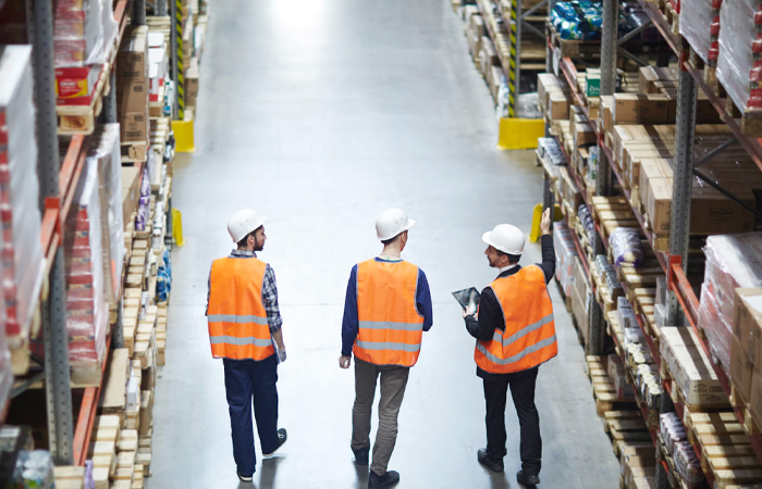 Three warehouse workers in safety gear discussing inventory management in a well-organized aisle,