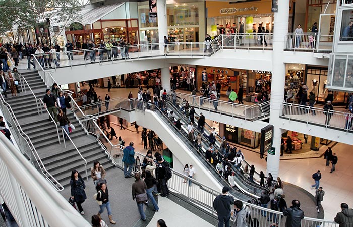 The image displays a bustling multi-level shopping mall filled with numerous shoppers. The mall