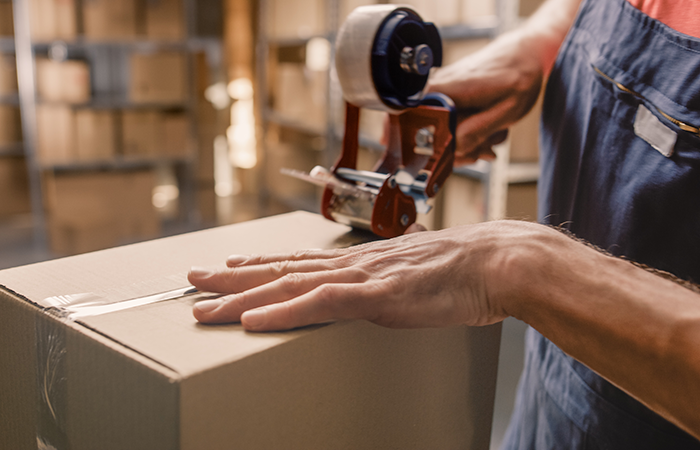 A warehouse employee in a blue apron is meticulously sealing a cardboard box with a