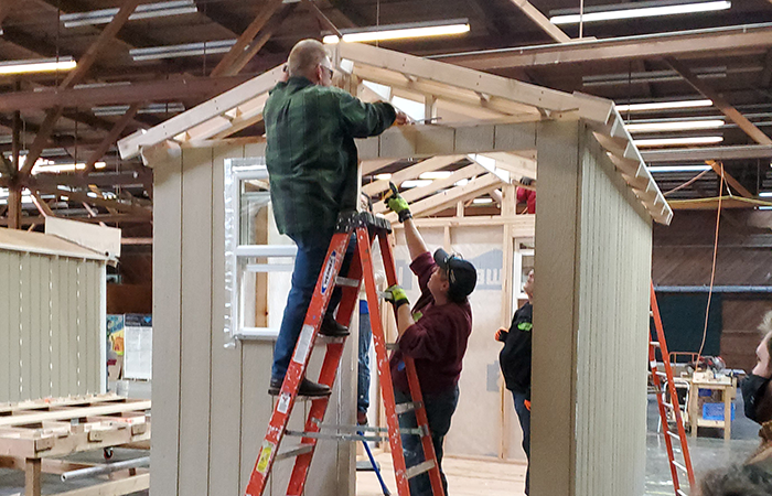 Two construction workers are engaged in building a wooden structure, with one on a ladder