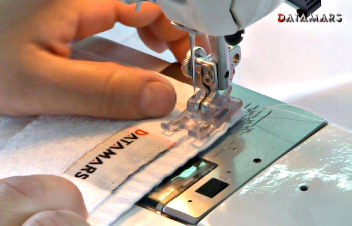 A close-up view of a sewing machine needle stitching a white RFID-enabled label with