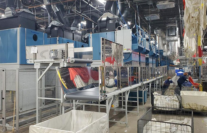 Inside a bustling industrial laundry facility, large blue commercial washing machines line the space,