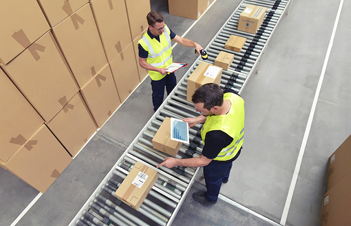 Two warehouse employees in safety vests are scanning and organizing packages on a conveyor belt system