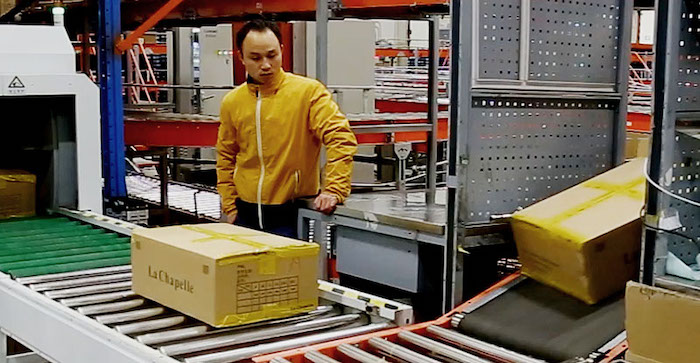 A worker in a yellow jacket stands beside a conveyor belt in a warehouse, monitoring