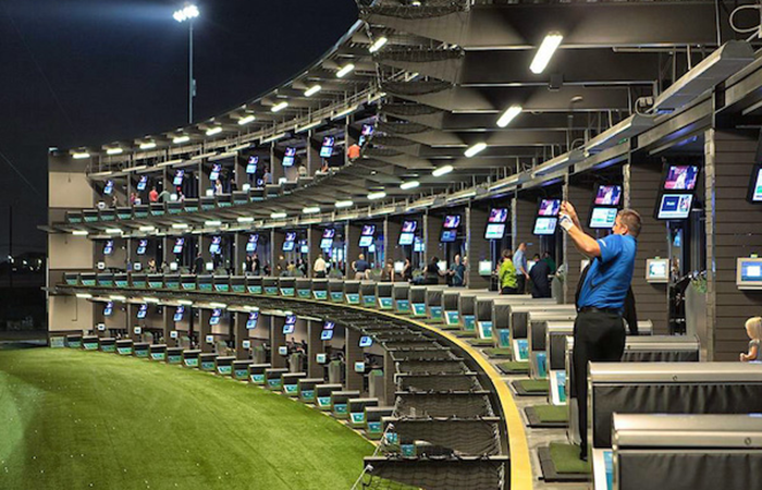 The image showcases a vibrant, modern driving range at night, illuminated by overhead lights