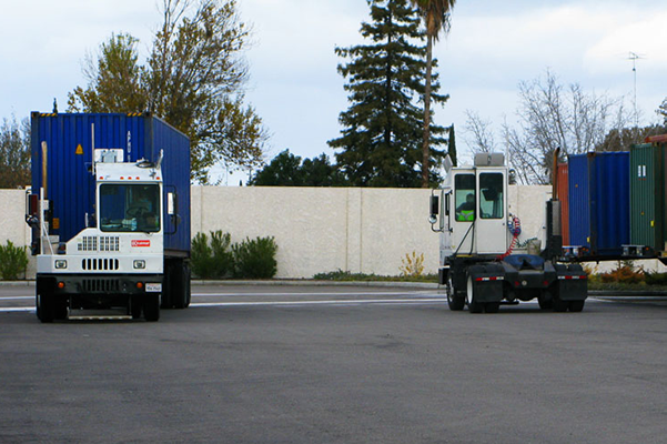 A commercial shipping yard with a white cab semi-truck transporting a blue shipping container