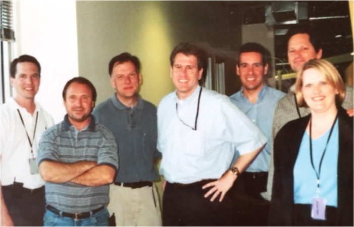 A group of professional individuals in casual and business attire stands together in an office setting